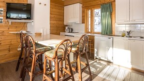 Une cuisine chaleureuse, aménagée dans le style d’un chalet, avec des murs en bois, une table à manger entourée de chaises, un téléviseur et une fenêtre agrémentée de rideaux.