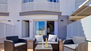 Un espace extérieur aménagé avec des meubles en osier et une table en verre, situé devant un bâtiment doté de balcons et de grandes fenêtres.