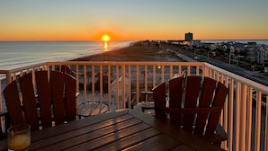 Une terrasse avec des chaises et une table en bois, offrant une vue sur une plage et l’océan au coucher du soleil.