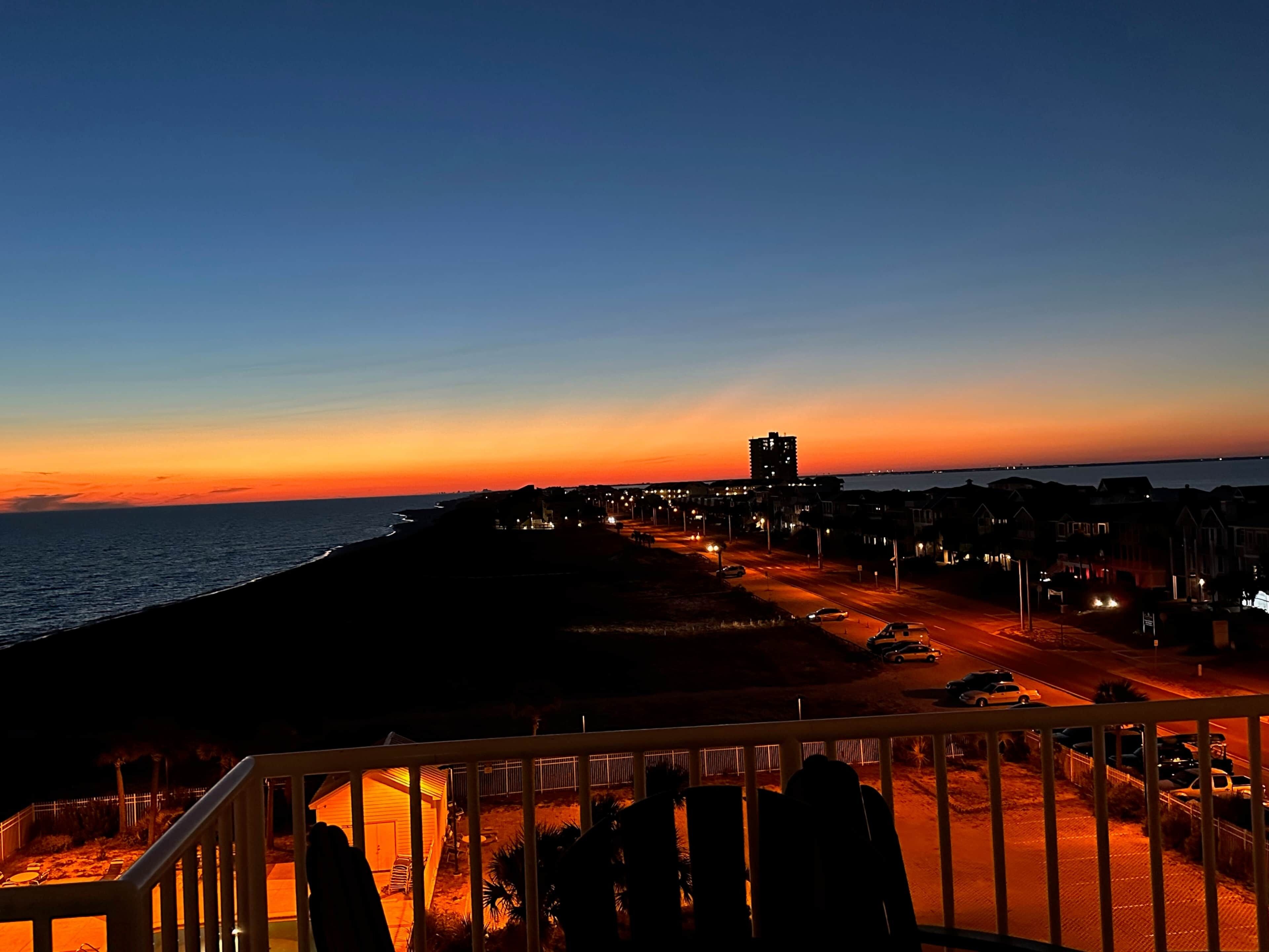 Un pueblo costero al atardecer, con un edificio destacado en la distancia, una calle con coches aparcados y vistas al mar.