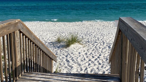 Une promenade en bois mène à une plage de sable fin bordée d’une eau d’un bleu limpide.