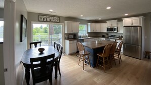 A spacious kitchen with white cabinets, a stainless steel refrigerator, and a dining area with wooden chairs. - Wine Trail, Cozy Cabin plus hot tub (Cobden)
