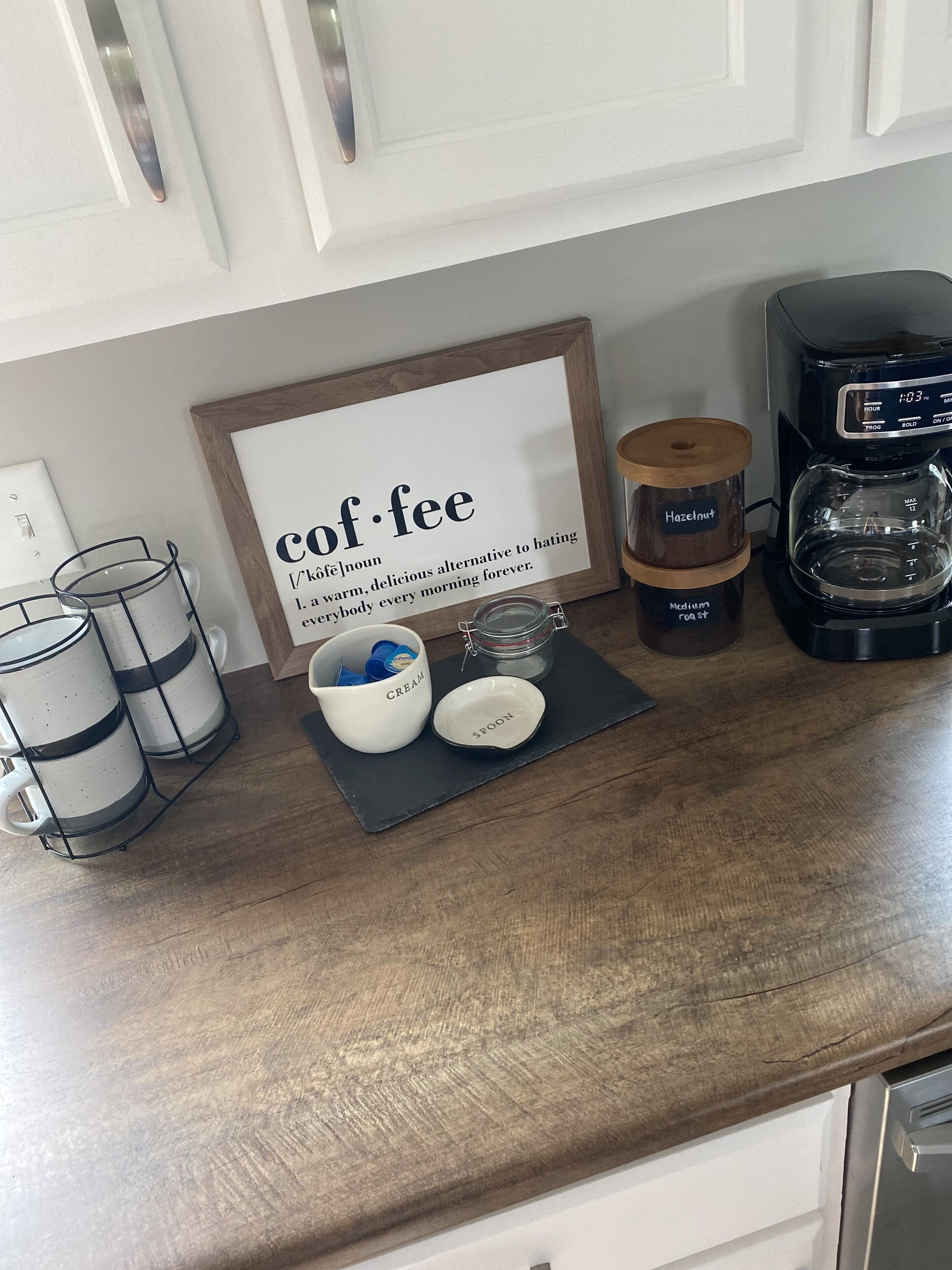 A kitchen counter with a coffee maker, a framed sign with the word 'coffee,' and a mug rack.