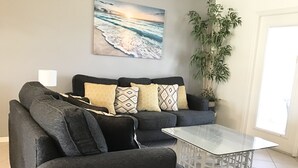 En moderne stue med en grå sofa, mønstrede puder, et glas-sofabord, en stor potteplante og et maleri af en strand på væggen.