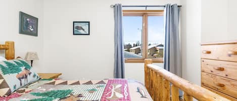 Un dormitorio con cama, cabecero de madera, cómoda, ventana con cortinas y vistas a casas.