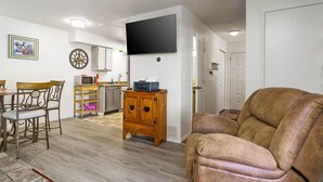 Un salon avec un canapé en cuir marron, un meuble en bois orné de découpes en forme de cœur, un téléviseur à écran plat fixé au mur et un coin cuisine équipé d’un four à micro-ondes et d’une étagère en bois.
