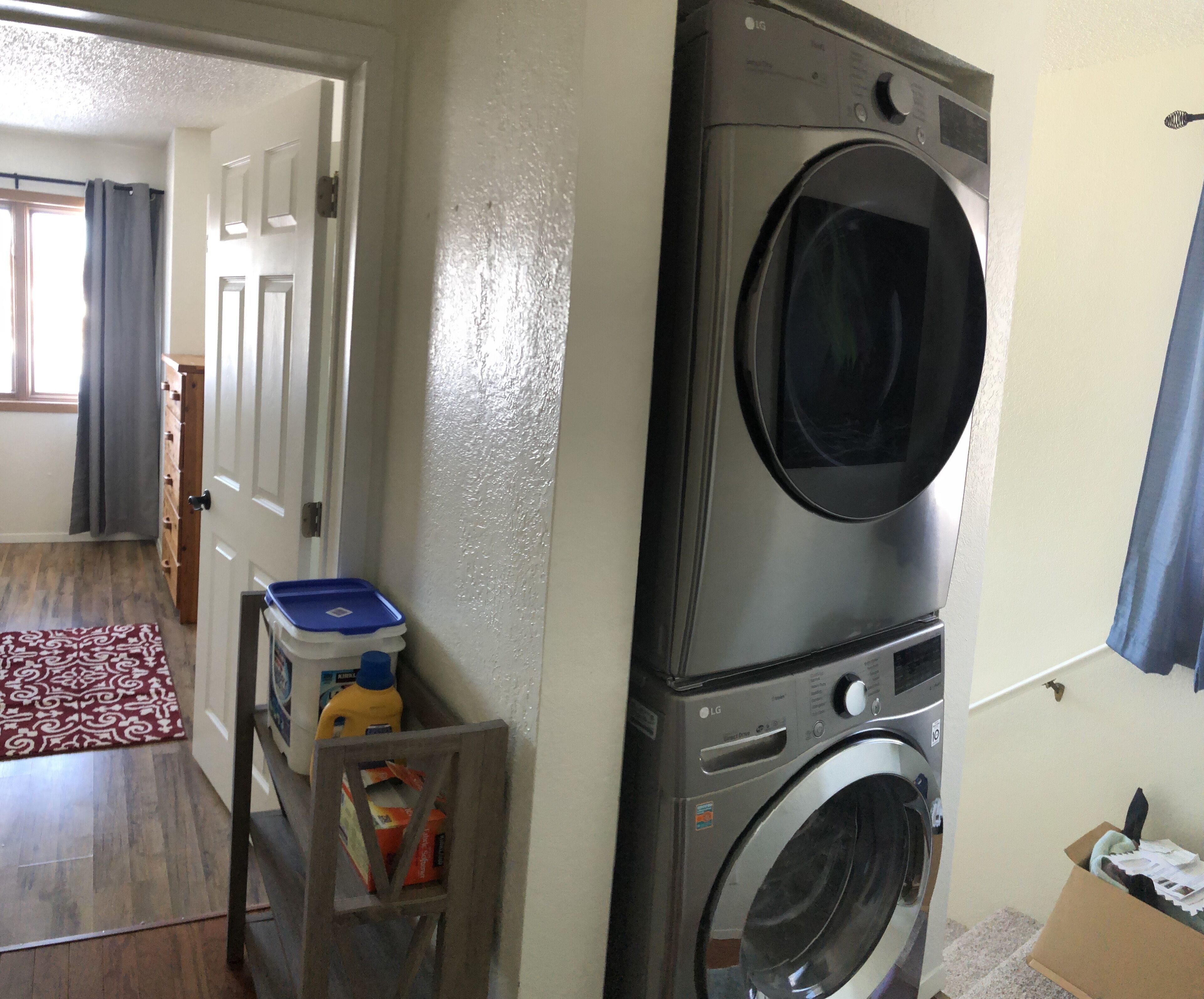 A laundry area with a front-loading washing machine, a drying rack, and a storage bin with cleaning supplies.