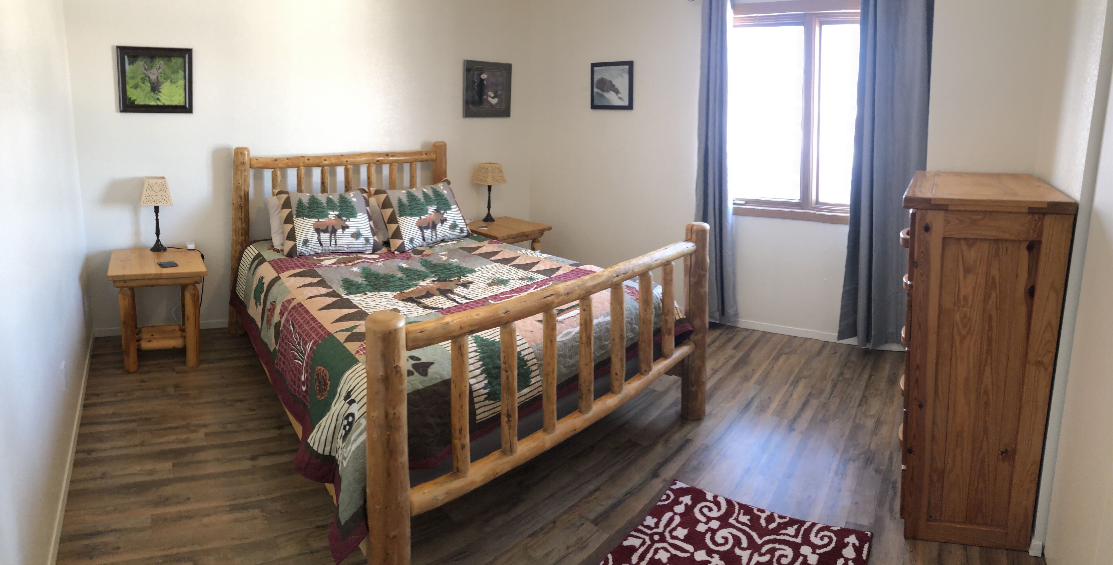 A wooden bed with a quilt, two bedside tables, a nightstand, a window with curtains, and a wooden cabinet.