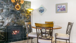 Une cheminée en pierre avec un feu de bois, une table ronde en bois entourée de chaises, et deux œuvres d’art encadrées accrochées au mur.