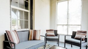 A covered outdoor seating area with wicker furniture and cushions, large windows, and a white brick wall. - Chic 2BR Stay | Cozy Living & High-Speed WiFi (Montgomery)