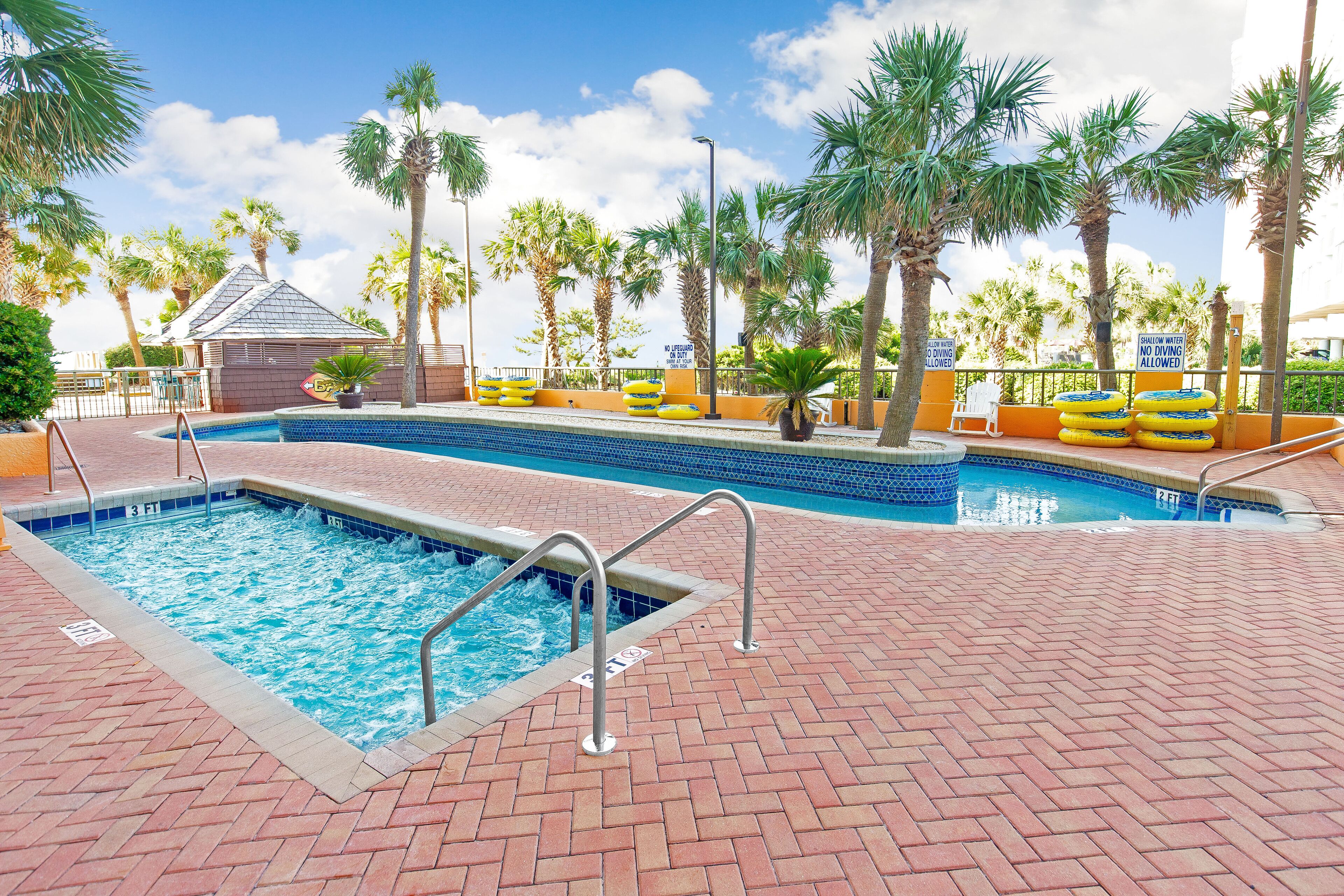 A swimming pool with a brick deck, surrounded by palm trees and a small building.