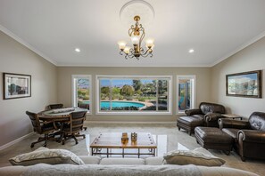 A spacious living room with a dining area, leather furniture, a chandelier, and large windows with a pool view.