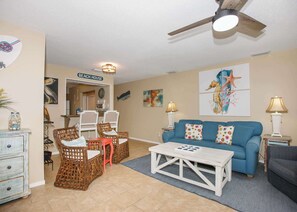 A beach-themed living room with wicker furniture, a blue sofa with colorful cushions, and a ceiling fan.