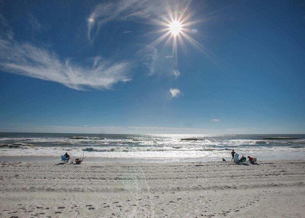 A beach with people sitting and walking along the shore, under a clear sky with the sun shining.