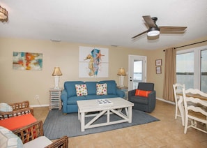 A living room with a blue sofa, a white coffee table, wicker chairs, and a ceiling fan.