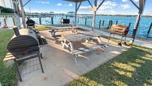 An outdoor covered patio with a grill, picnic table, and seating area overlooking a body of water.