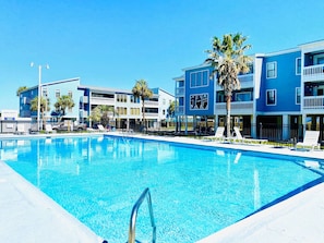 A clear blue swimming pool with lounge chairs and palm trees, surrounded by multi-story buildings with balconies.