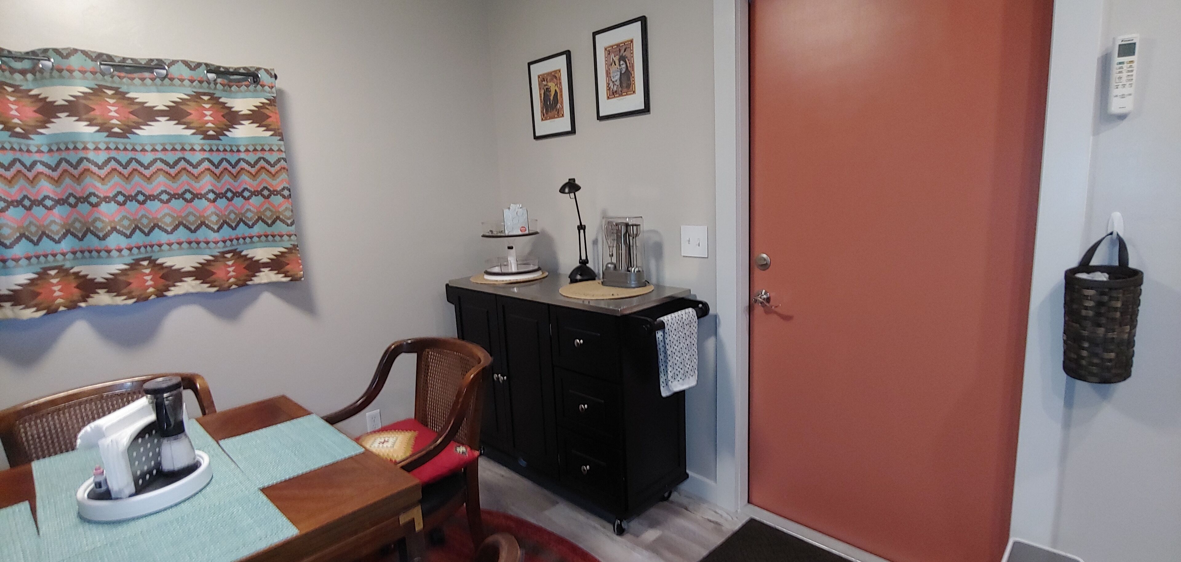 A dining area with a wooden table, chairs, a coffee maker, a black cabinet with a towel hanging, and a red door.