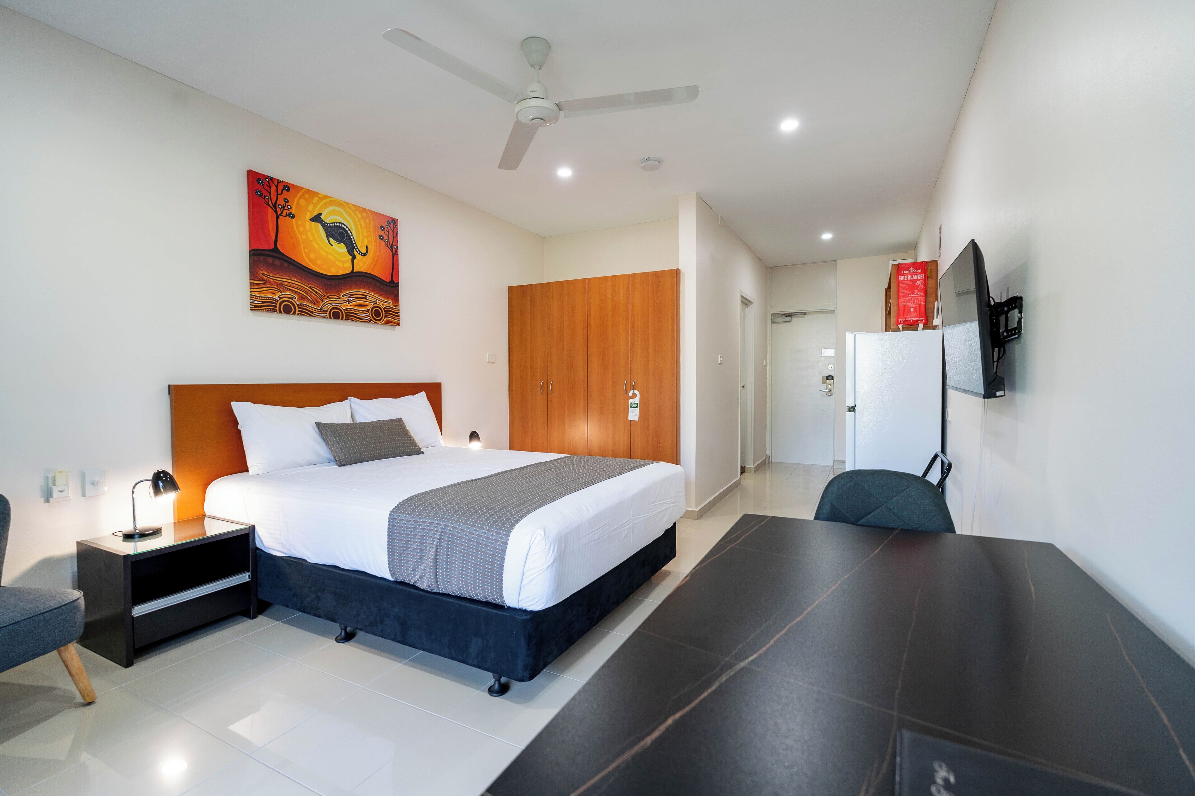 A modern hotel room with a large bed, a desk, a chair, a ceiling fan, and a painting on the wall.
