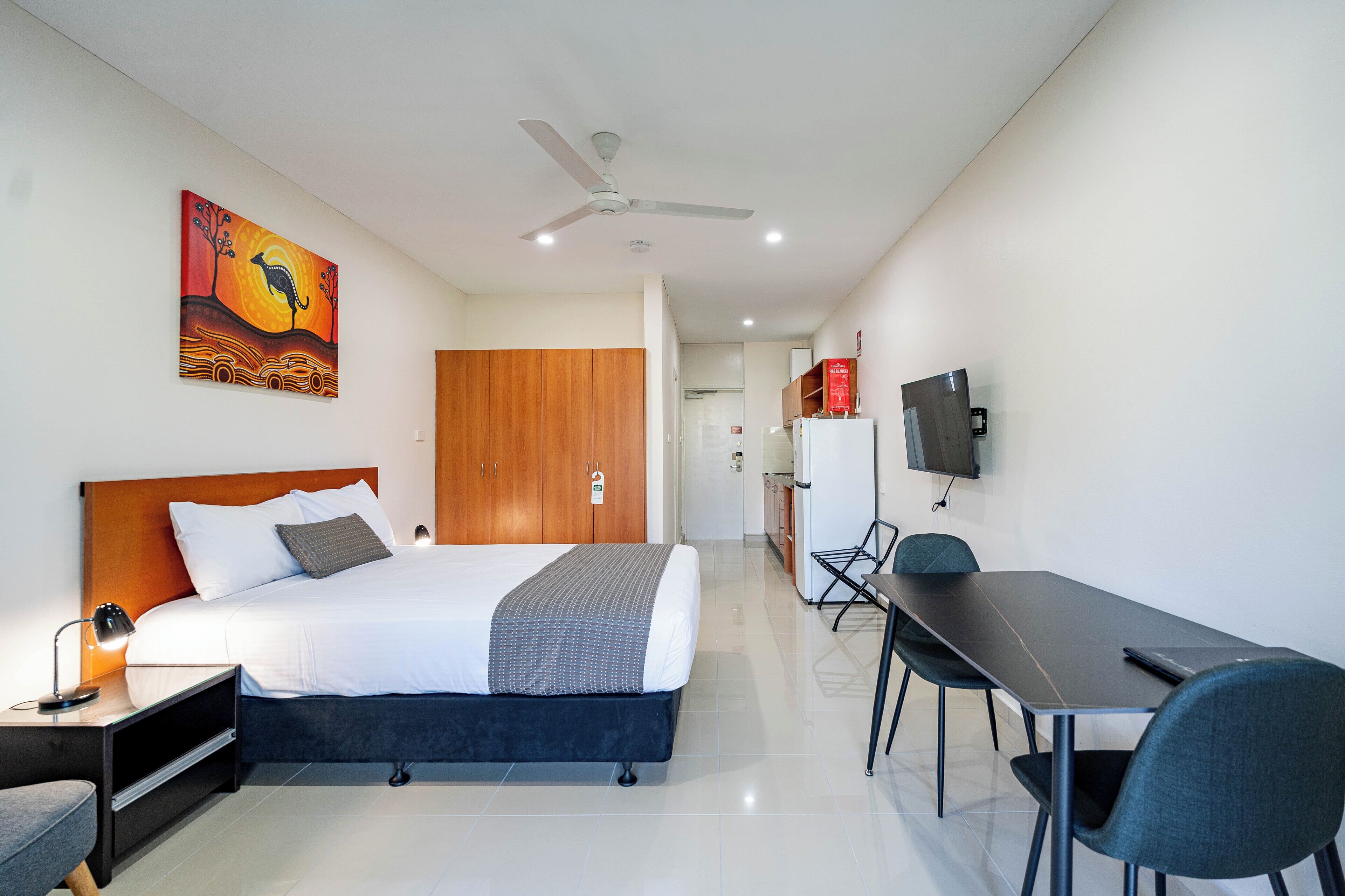 A hotel room with a bed, desk, chairs, a TV, and a painting on the wall.