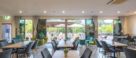 Un vaste espace intérieur aménagé pour les repas, avec de nombreuses tables et chaises, de grandes fenêtres dotées de rideaux et une vue sur la terrasse extérieure.