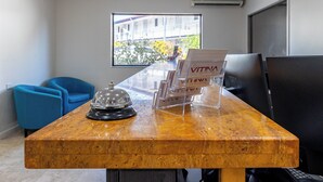 Un mostrador de recepción con encimera de madera, un timbre y un atril para revistas. Hay sillas azules y un ventanal con vistas a edificios y árboles.