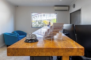 A reception desk with a wooden top, a bell, and a magazine rack. There are blue chairs and a large window with a view of buildings and trees. - Vitina Studios by Argus (Darwin)