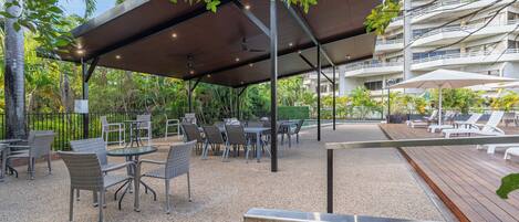 An outdoor patio area with tables and chairs, a covered seating area, and a view of a multi-story building in the background.