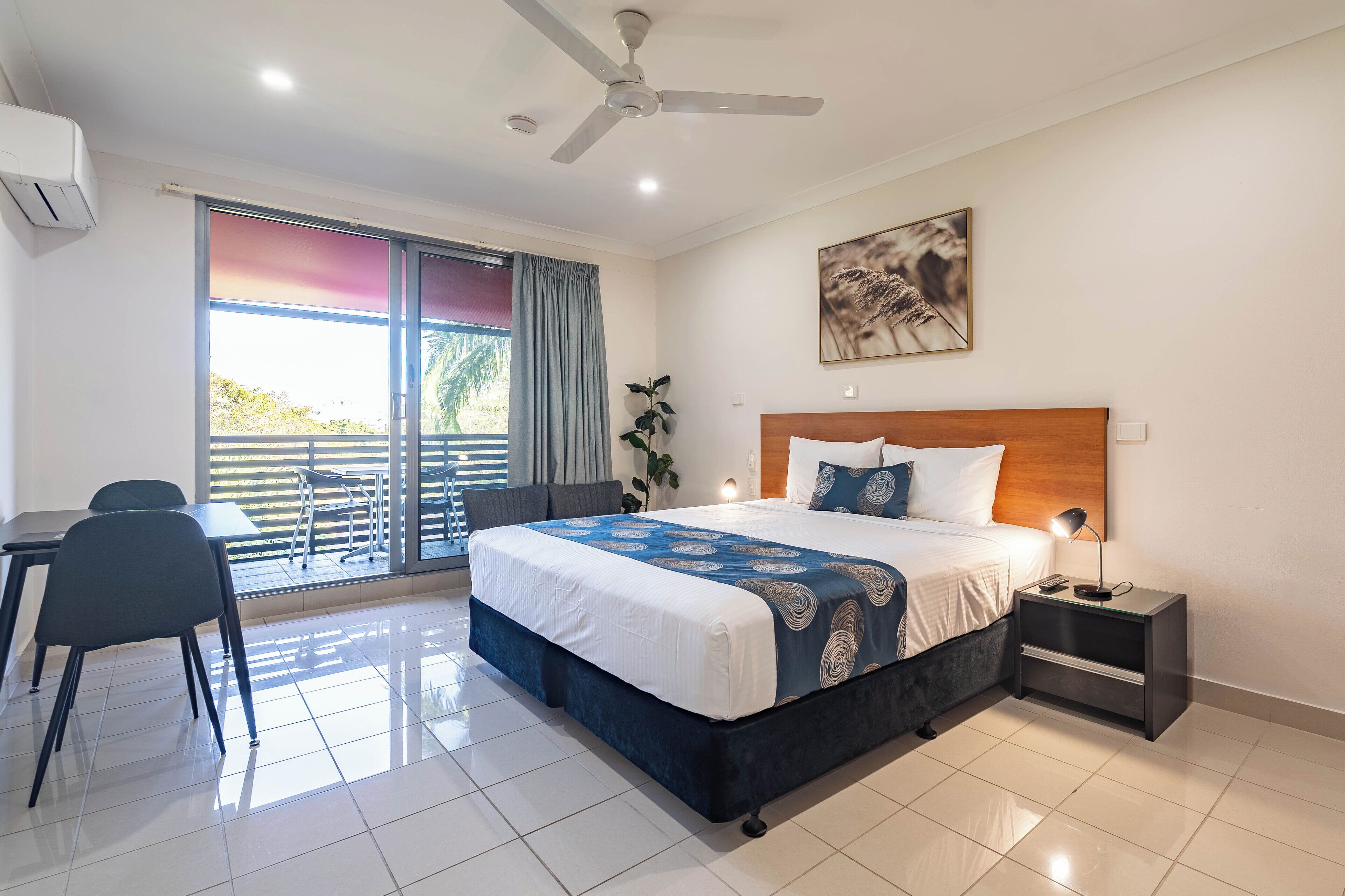 A hotel room with a bed, a desk, a chair, a nightstand, a ceiling fan, and a large window with curtains.