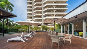 Un terrazza in legno con poltrone da relax e ombrelloni, sullo sfondo un grattacielo e l'ingresso con una porta in vetro.