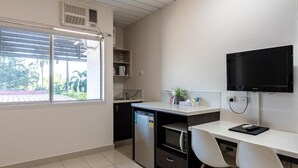 Una cocina compacta con microondas, refrigerador y un lavamanos pequeño. Hay un escritorio blanco con una silla y un televisor montado en la pared.