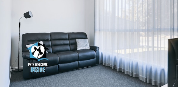 A black leather sofa with a cushion, a floor lamp, sheer curtains, and a television on a stand. - 32 Tabernacle Rd, Encounter Bay - Brand New 3BR So Close to Sea and Lake (Encounter Bay)