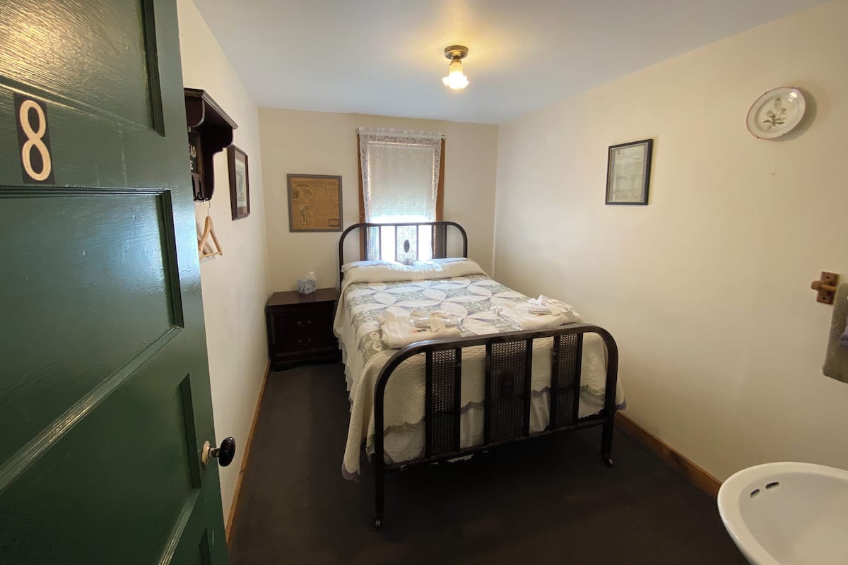 A small, clean room with a single bed, a bedside table, a wall-mounted toilet, and a door with a number.