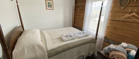 Una habitación pequeña y acogedora con una cama individual, un cabecero de madera, una mesita con toallas y una ventana con cortinas de encaje.