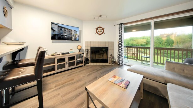 A modern living room with a fireplace, a television, and a balcony with a view of greenery. - Aurora at Nautilus (Ocean Shores)