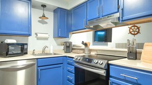 A kitchen with blue cabinets, stainless steel appliances, a coffee maker, and a ship's wheel decoration. - Aurora at Nautilus (Ocean Shores)