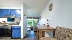 A compact kitchen with blue cabinets, a dining area with a wooden table and woven sofa, and a view of a green garden through a glass door. - Aurora at Nautilus (Ocean Shores)