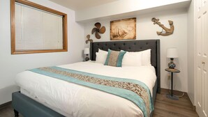 A neatly made bed with a headboard, a window with blinds, and decorative wall art including an anchor and a sailboat painting. - Aurora at Nautilus (Ocean Shores)