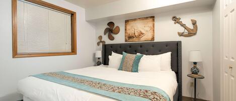 Una cama bien hecha con cabecera, una ventana con persianas y decoración en la pared, incluyendo un ancla y un cuadro de un velero.