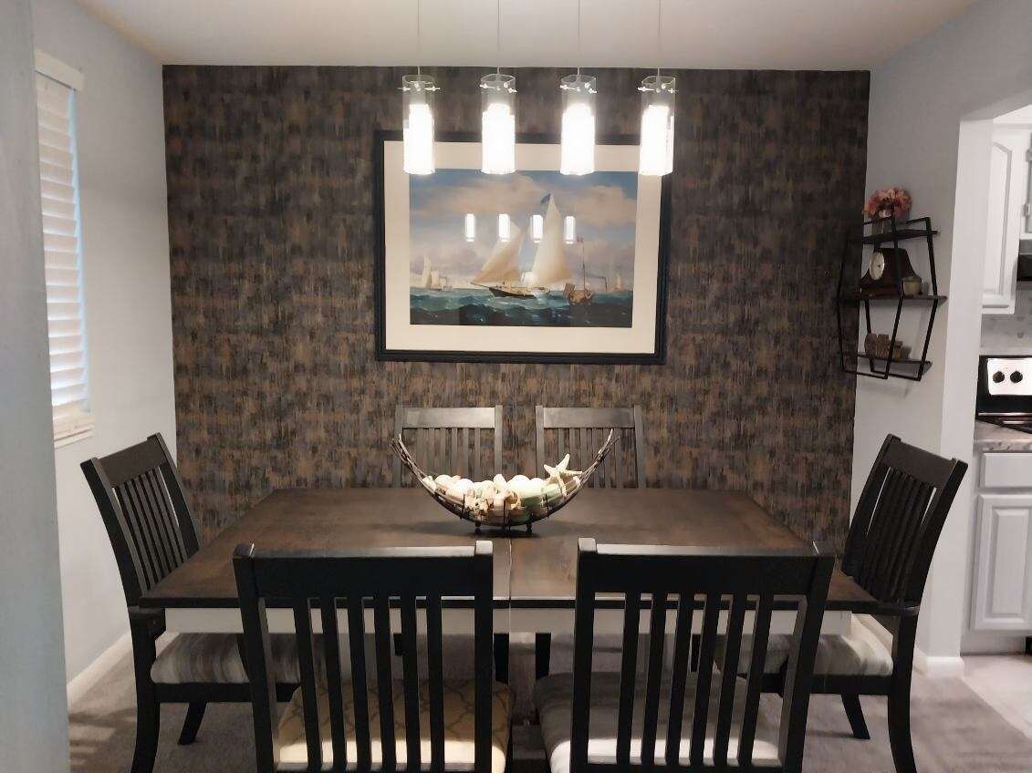 Un coin repas avec une table en bois foncé, quatre chaises, un grand saladier au centre et un tableau encadré représentant des voiliers accroché au mur.