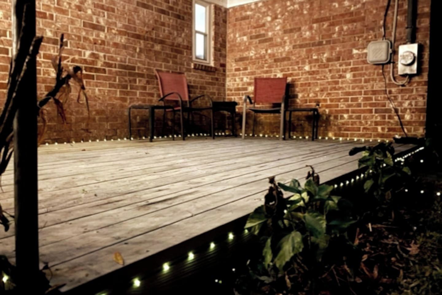 Una terraza de madera con sillas, un muro de ladrillo y una ventana.