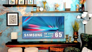Een woonkamer met een grote Samsung SmartTV op een houten standaard, een lijst met een woordwolk aan de muur en decoratieve objecten op planken.