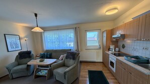 Un espace de vie compact comprenant un canapé, des fauteuils, une petite table, un radiateur, une fenêtre avec des rideaux, une kitchenette avec des placards en bois et un luminaire au plafond.