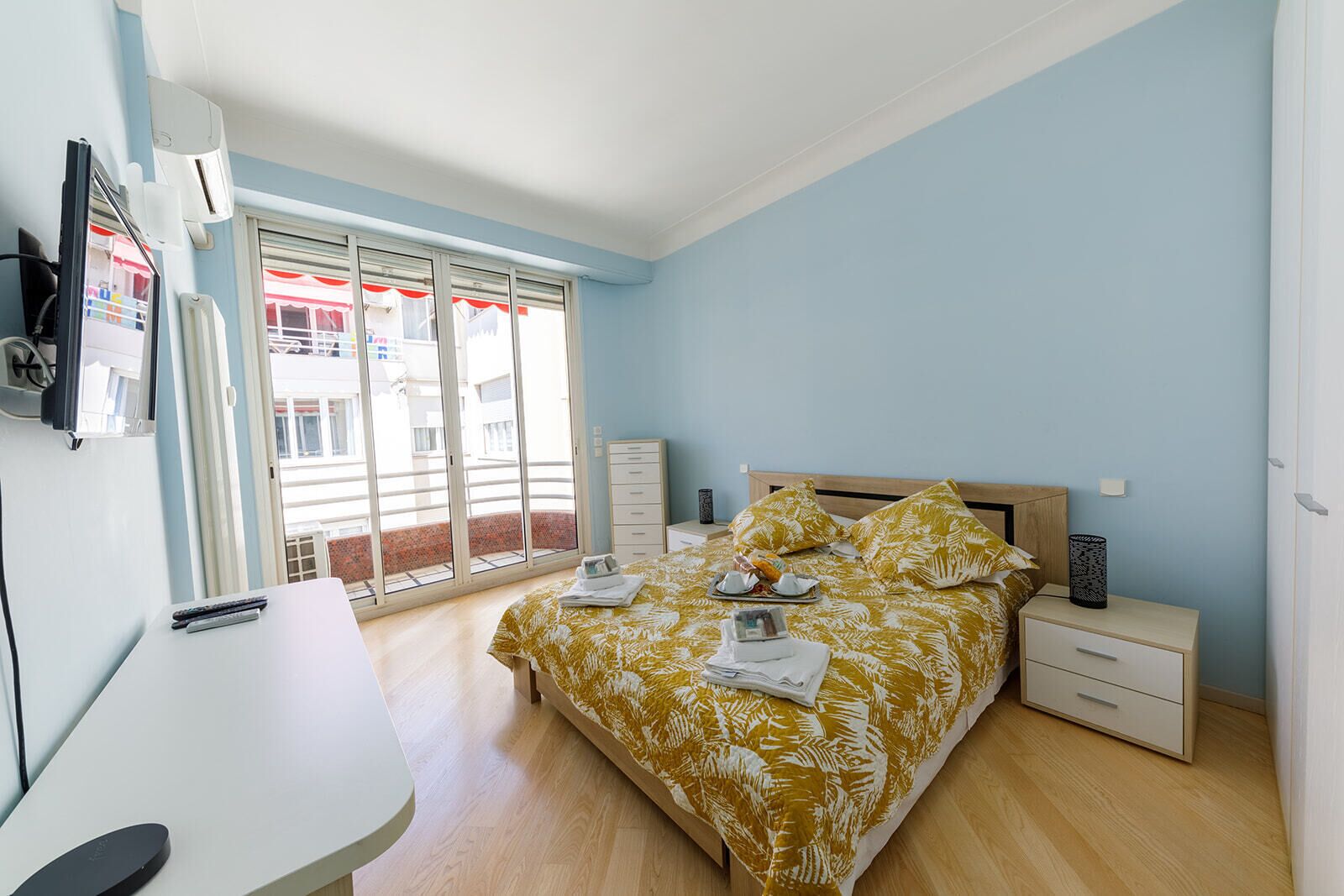 A bedroom with a bed, a desk, a chair, a nightstand, and a window with a view of buildings.