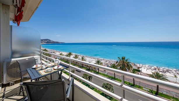 A balcony with a table set for tea, overlooking a beach with palm trees and a clear blue sky. - Wonderful Apartment on Sea (Nice)
