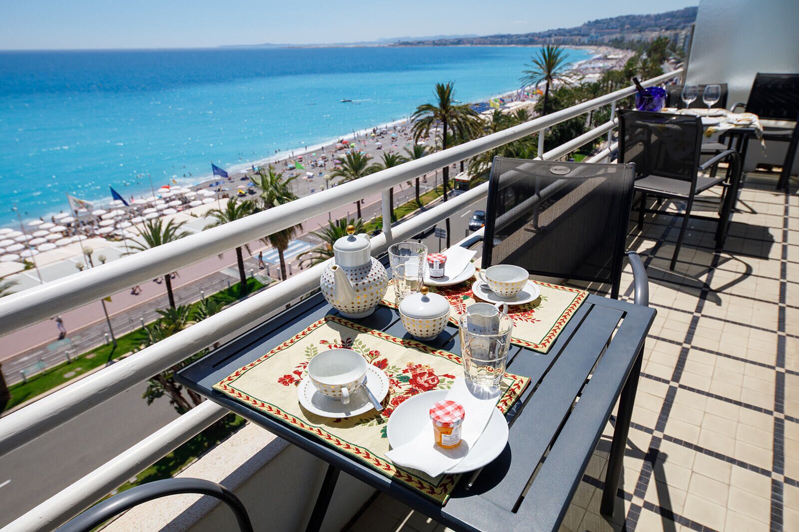A balcony table set with tea set and glasses, overlooking a beach and cityscape.