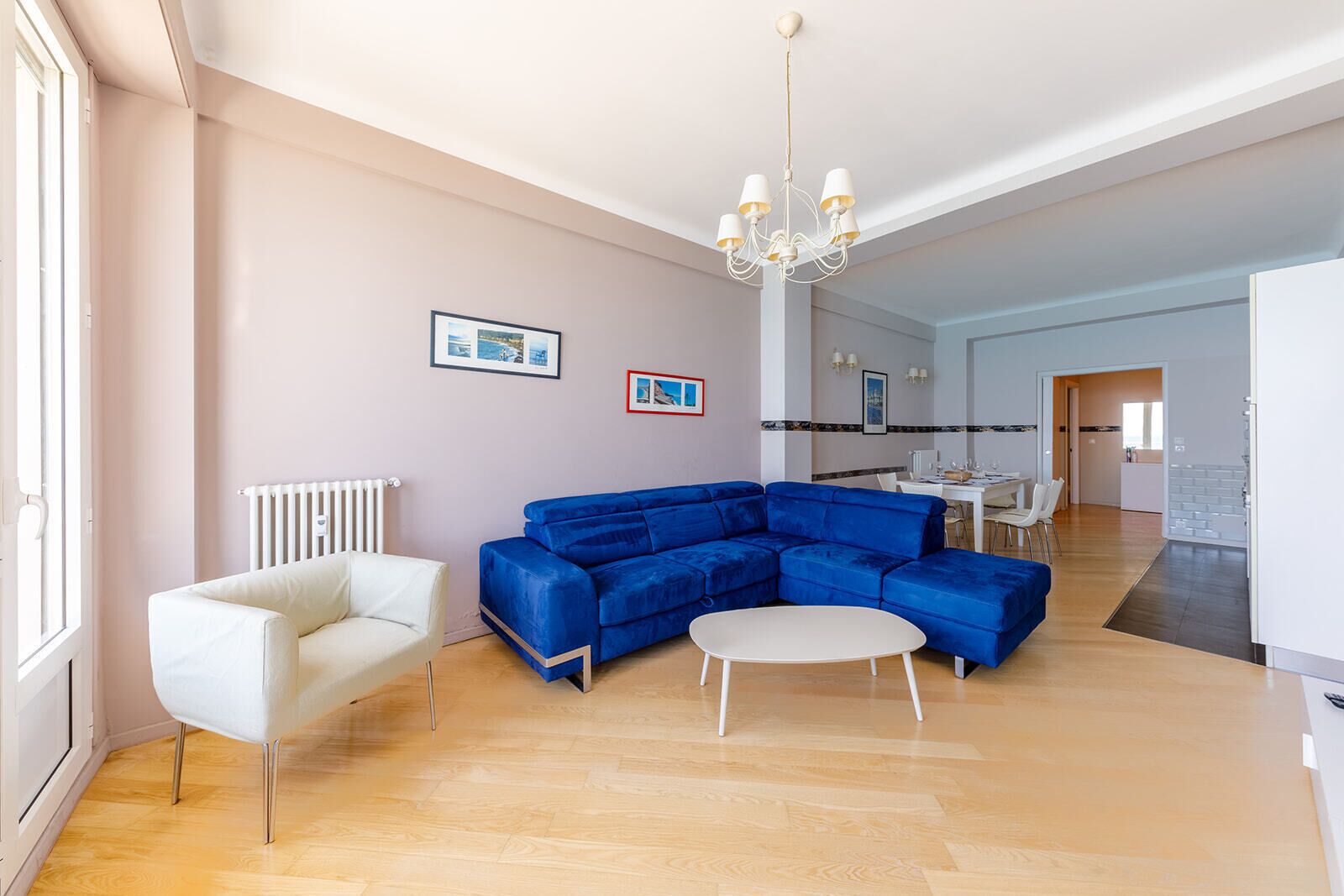 A modern living room with a blue sectional sofa, a white armchair, and a round coffee table. There is a chandelier, a dining area in the background, and a kitchen visible through an open door.