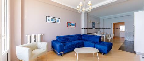 A modern living room with a blue sectional sofa, a white armchair, and a round coffee table. There is a chandelier, a dining area in the background, and a kitchen visible through an open door.