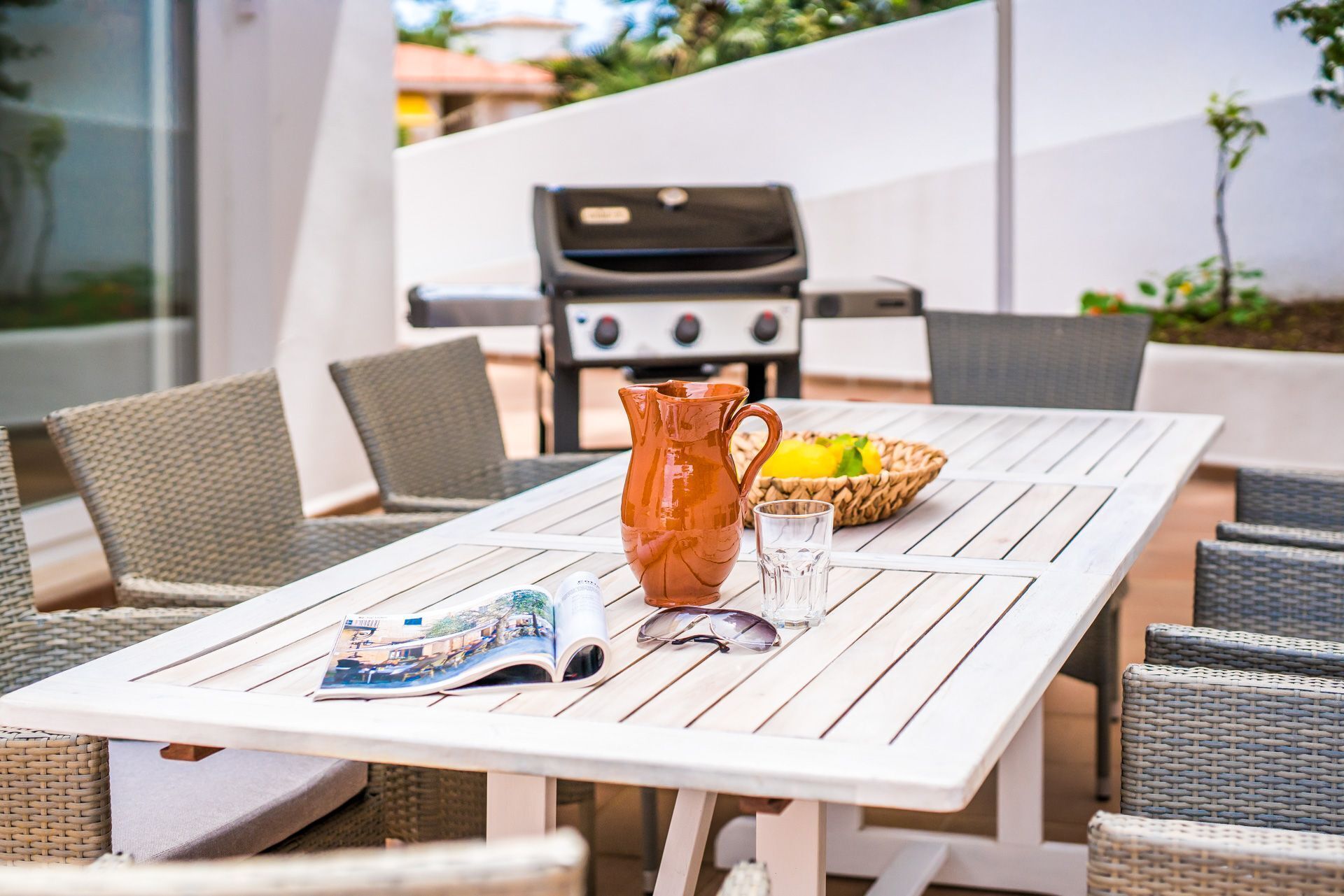 Un patio con mesa blanca, sillas de mimbre, una parrilla y una cesta de fruta.