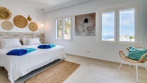 Un dormitorio con cama, una silla de mimbre y vista al mar a través de amplias ventanas.